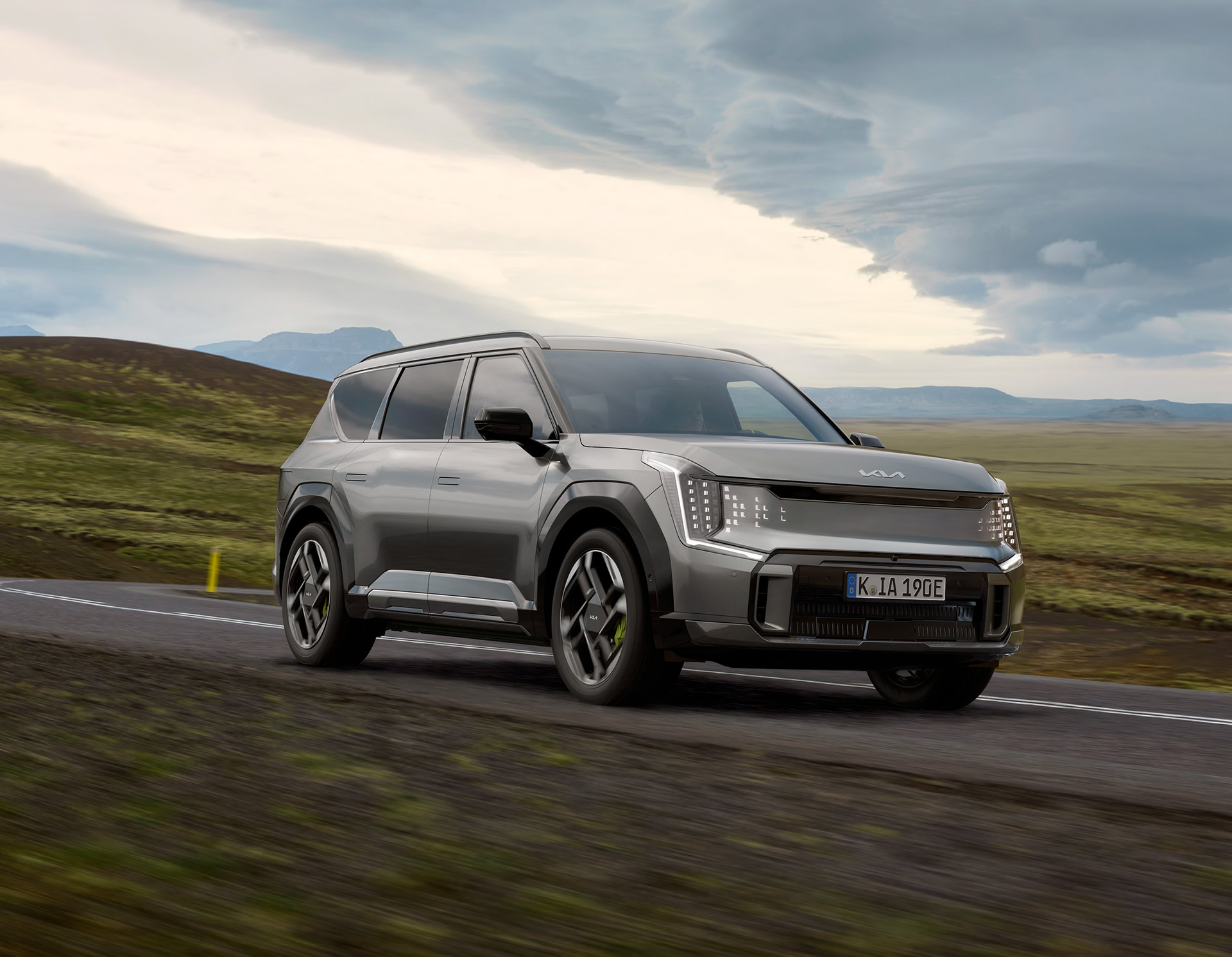 Kia EV9 GT, grau, seitliche Frontansicht, Beifahrerseite, Fahrzeug fährt durch Landschaft